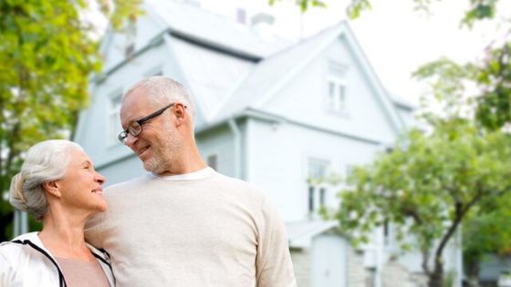 Elderly couple smiling at each other in front of their home surrounded by greenery on a sunny day.