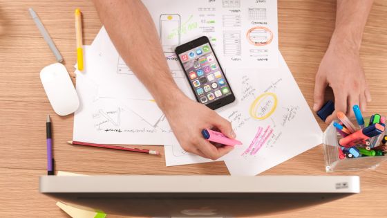 Person planning app design with documents, smartphone, and desktop monitor on desk, highlighting with markers.