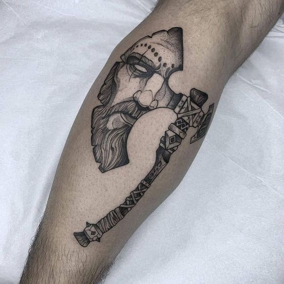 Nordic warrior tattoo on calf with detailed helmet and beard, combined with an ornamental axe, showcasing intricate line art.