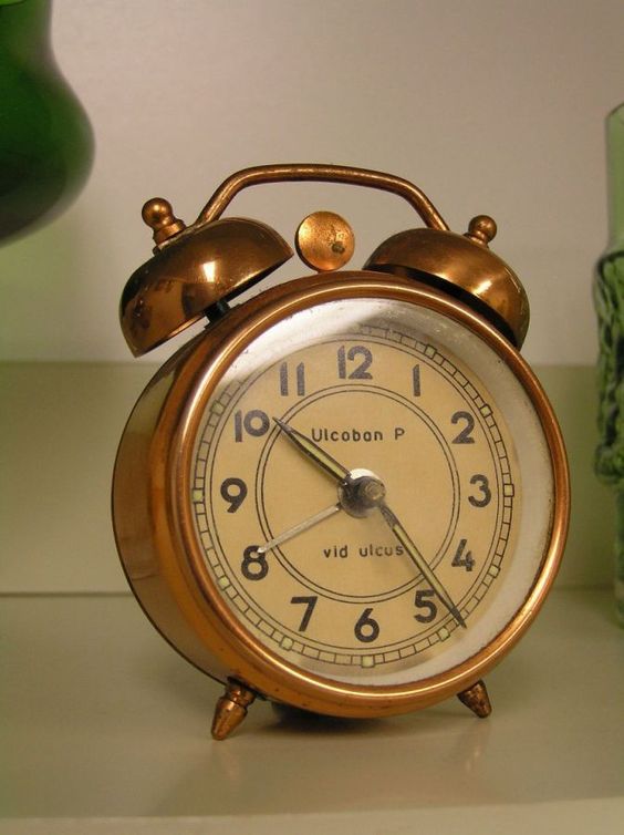 Vintage brass alarm clock with a beige face and black numerals, set on a shelf with green decor elements.