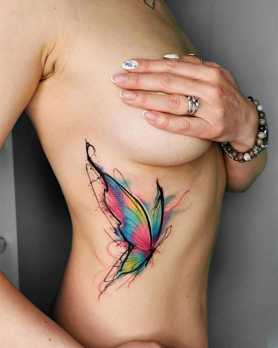 Colorful butterfly tattoo on a woman's side rib cage, featuring vibrant hues of blue, pink, and yellow with abstract outlines.