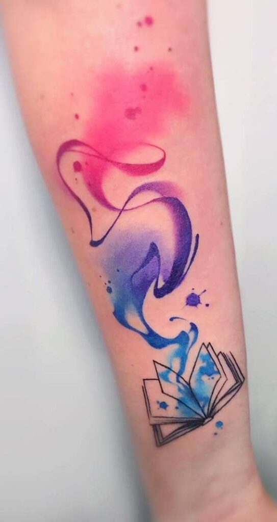 Forearm tattoo of an open book with colorful watercolor ink splashes and swirling designs in blue, purple, and pink shades.