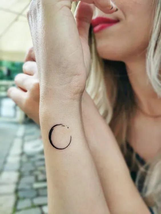 Minimalist black ink crescent moon tattoo on a person's forearm, with a blurred background and bright daylight.