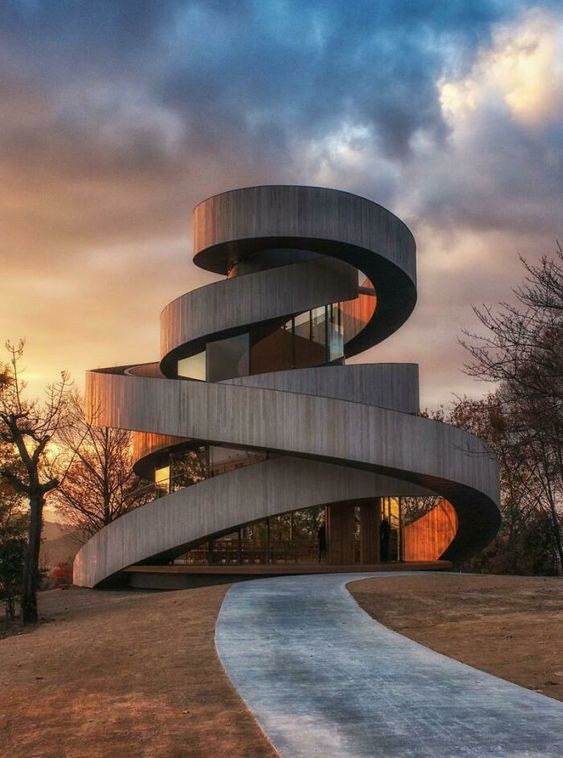 Modern spiral architectural building with sleek design at sunset, with a winding path leading to the entrance.