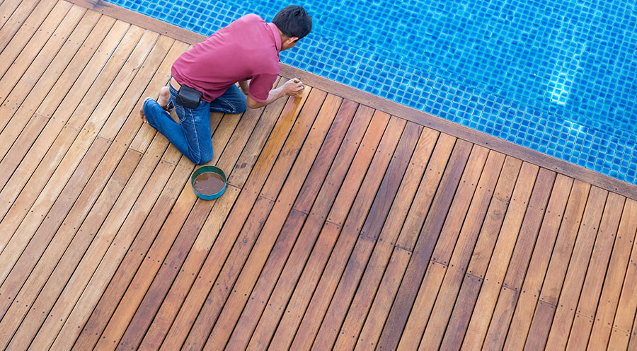 Man kneeling and varnishing a wooden deck beside a blue tiled pool, enhancing outdoor space by pool maintenance and care.