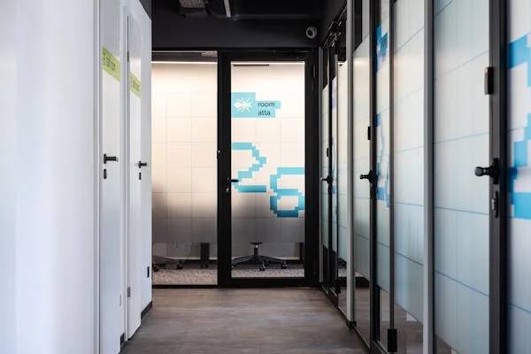 Modern office hallway with glass doors leading to conference room labeled Room 25 in blue pixel design.