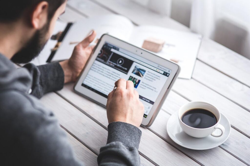 Person browsing website on tablet with stylus, while having coffee; modern work setup on a wooden desk.