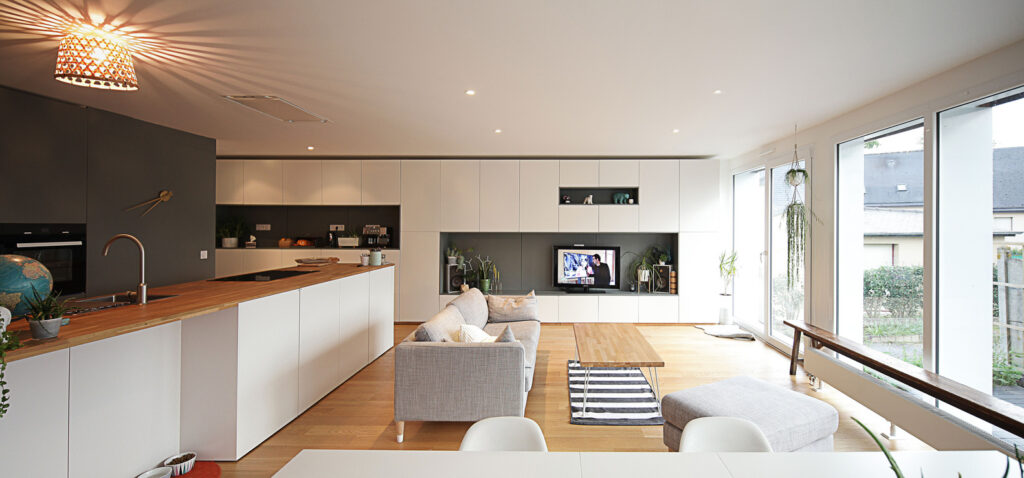 Modern open-plan living room and kitchen with minimalist décor, large windows, and light wood flooring.