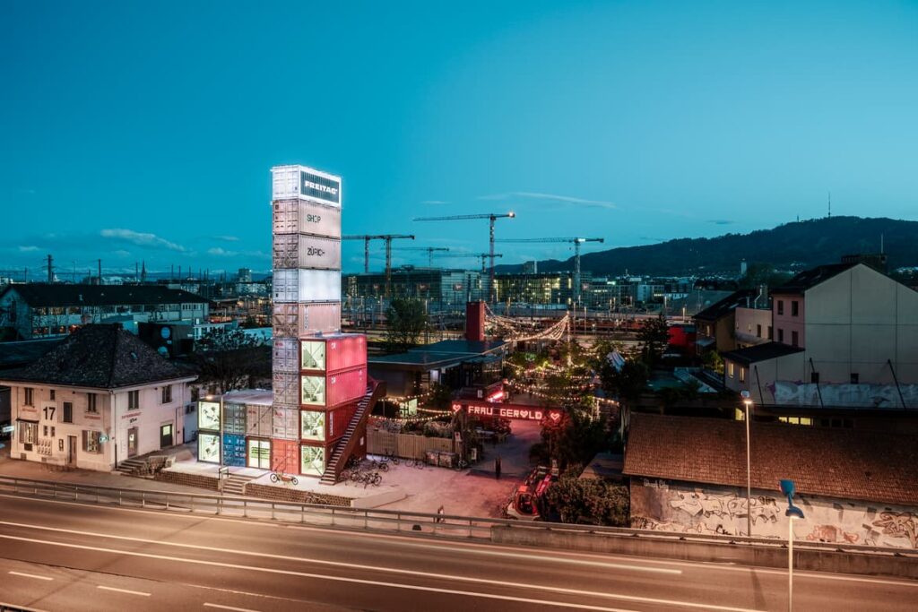 Freitag Tower and Frau Gerolds Garten in Zurich at dusk, vibrant lights highlighting the unique urban landscape and creative design.