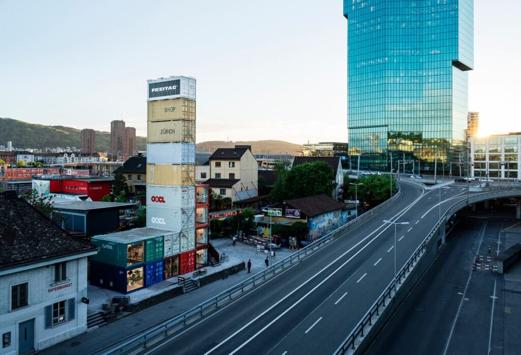 Stacked shipping containers next to a modern glass tower and a road in an urban setting with mountains in the background.