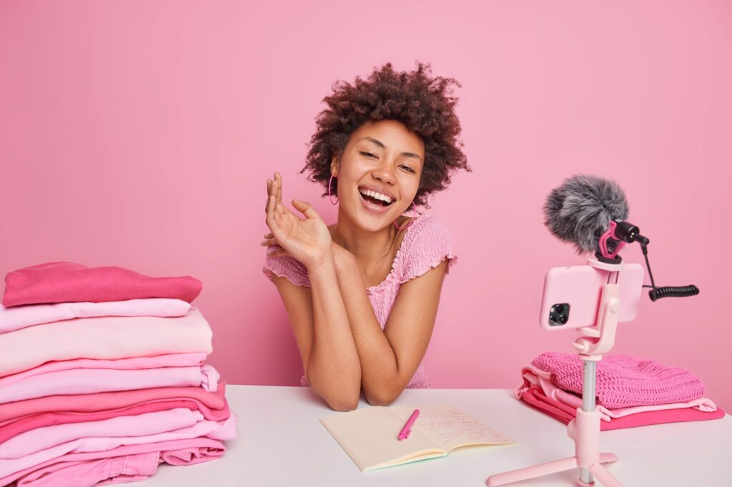 Smiling woman recording a vlog with pink clothes and notebook, set up in a home studio with pink background.