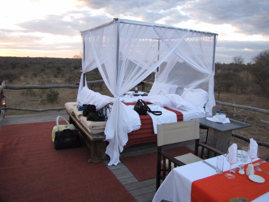 Outdoor bed with canopy and mosquito netting, overlooking a scenic landscape with a set dining table at sunset.