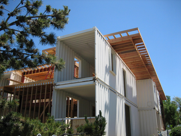 Shipping container house under construction, surrounded by trees, with a clear blue sky in the background.