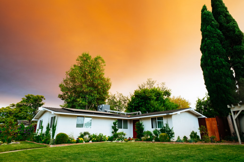 Single-story white house with a lush front yard and tall trees at sunset. Orange sky creates a serene and picturesque scene.