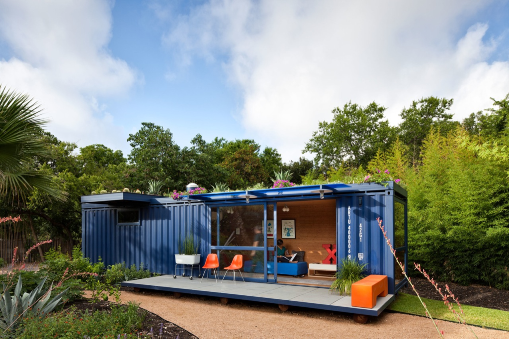 Modern cozy tiny house made from a blue shipping container amidst lush greenery and garden, outdoor sitting area visible.