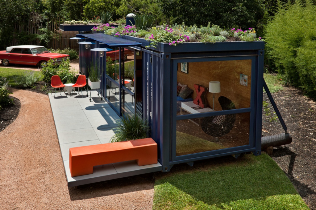 Modern shipping container house with rooftop garden, patio seating, and vintage red car in the lush green backyard.