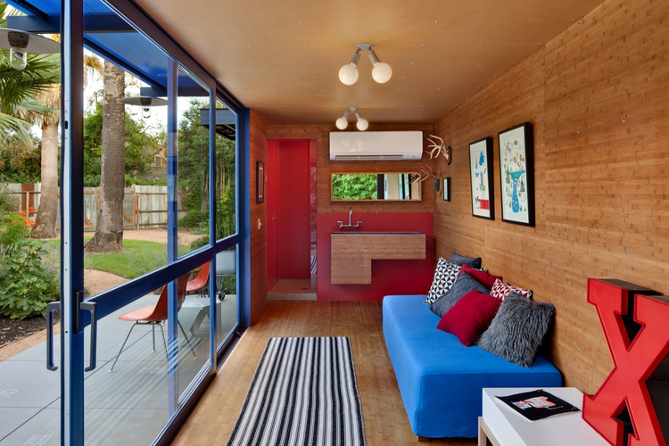 Modern tiny home interior with blue sofa, striped rug, wall art, and kitchenette. Large glass doors open to lush garden.