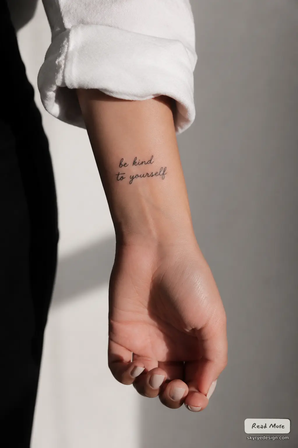 Minimal script wrist tattoo reading'be kind to yourself' on inner wrist, close-up of hand with rolled white sleeve