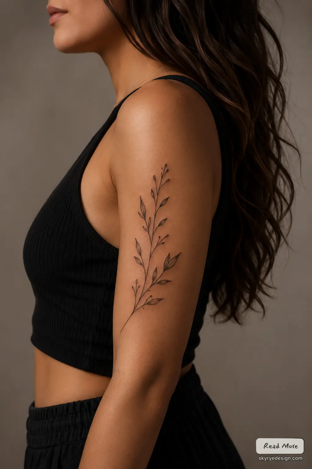 Upper arm botanical tattoo, minimal fine-line branch and leaves in black ink on woman wearing black crop top