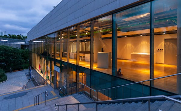 Modern building with glass windows at dusk, displaying interior lights and a staircase leading to the entrance.