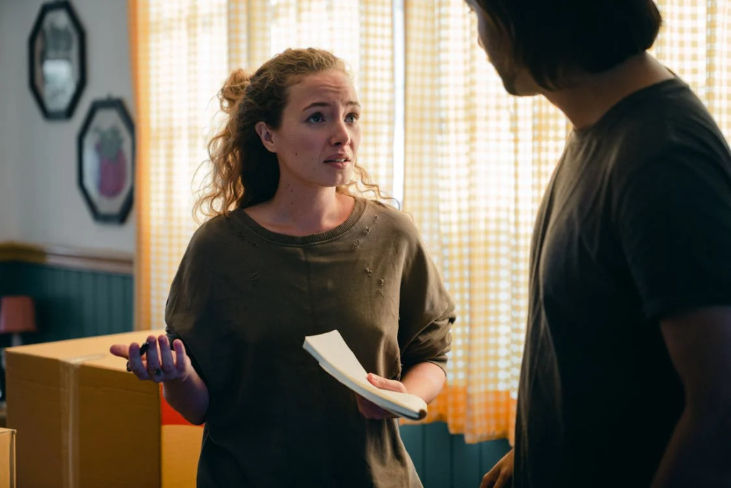 Woman holding a notepad, conversing with a man near moving boxes in a sunlit room with checkered curtains.