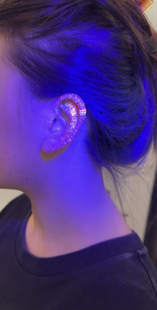 Close-up of a woman's ear wearing a sparkling, glittery ear wrap under blue lighting. Unique jewelry, glowing effect.