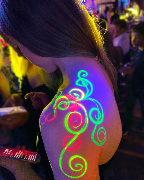 Close-up of a woman's shoulder with glowing neon body paint in a vibrant, colorful nightlife party scene.