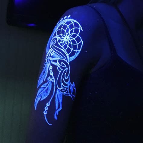 Glow-in-the-dark dreamcatcher tattoo on upper arm, featuring intricate feathers and geometric patterns under blacklight.