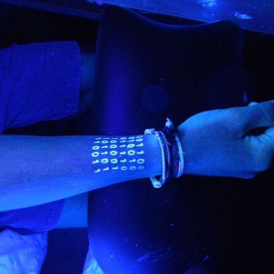 Arm under UV light with glowing binary code tattoo and braided bracelets, creating a sci-fi futuristic look.