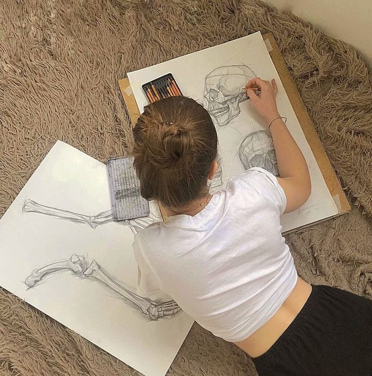 Top-down view of a person drawing anatomical skull and leg bone sketches on paper with pencils.