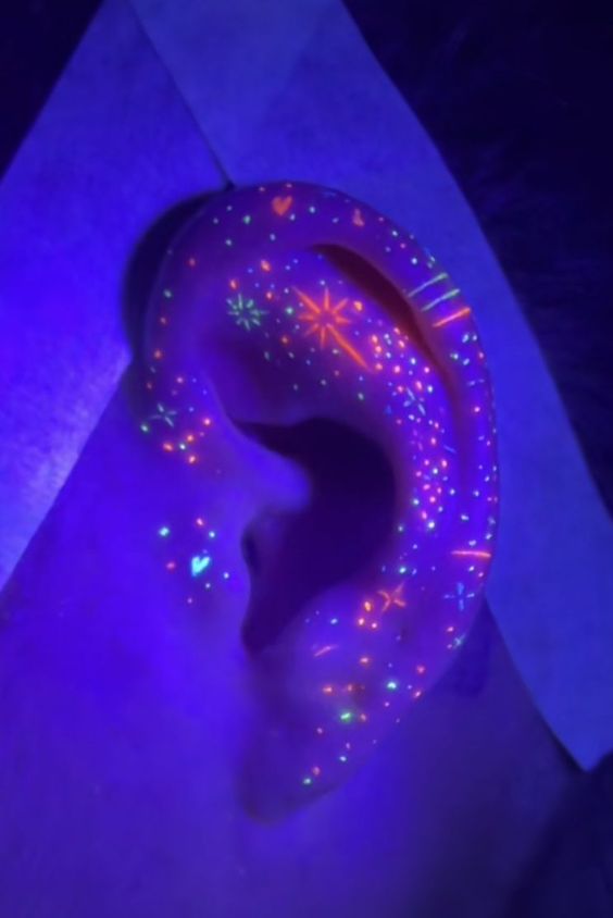 Close-up of an ear with glowing neon tattoo designs under ultraviolet blacklight, showcasing star and heart patterns.