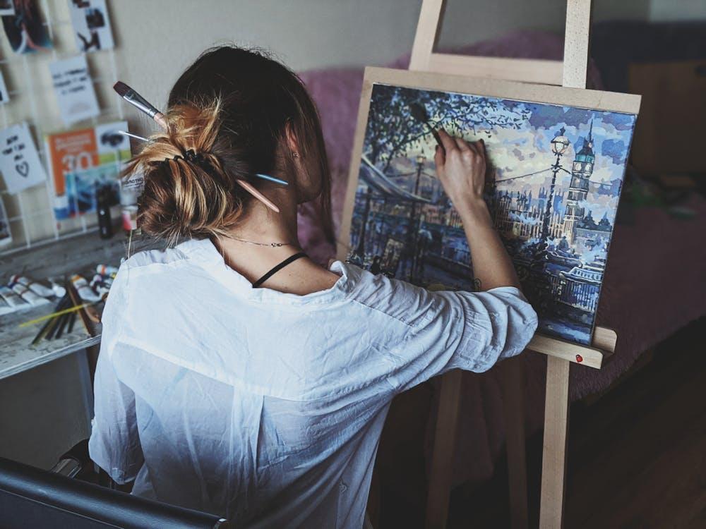 Artist painting vibrant cityscape on canvas at easel, tools and brushes in background. Creative studio atmosphere.