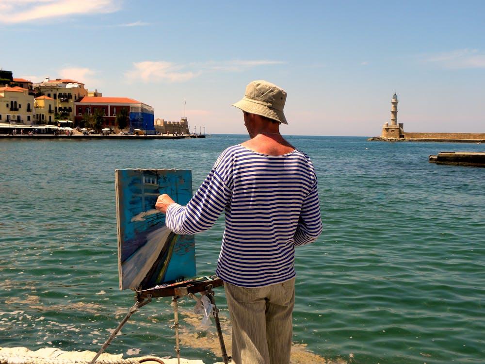 Artist painting seaside landscape with lighthouse and colorful buildings in background on sunny day.