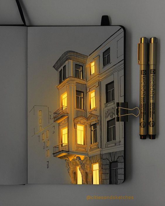 Sketch of a lit-up building on a notebook with glowing windows, next to two gold pens and a binder clip.