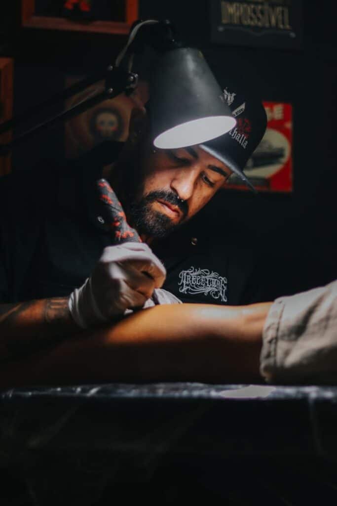 _ 8 Tattoo artist concentrating on creating intricate design on client's arm under focused lighting in a studio setting. | Sky Rye Design Tattoo artist concentrating on creating intricate design on client's arm under focused lighting in a studio setting.