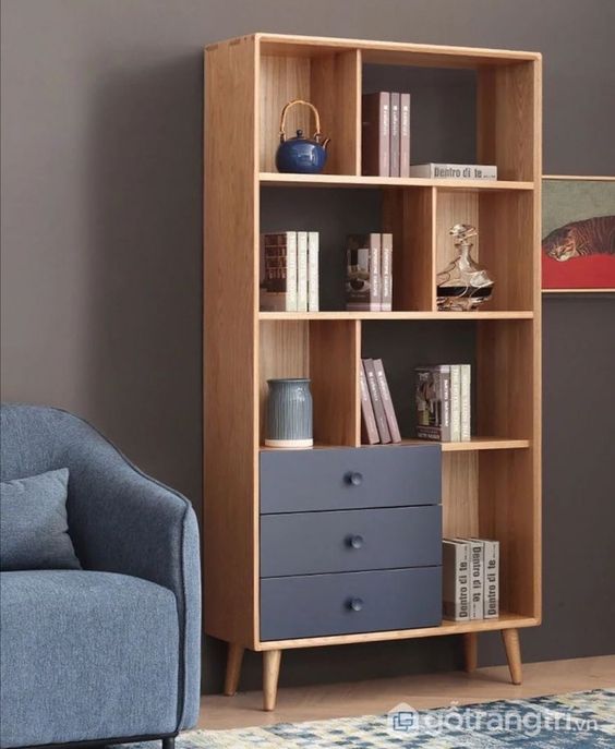 Minimalist wooden bookshelf with drawers and decor items next to a blue armchair against a dark wall.