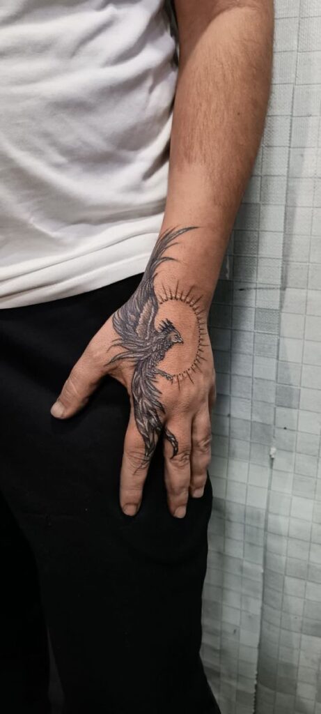 Anka Hand tattoo featuring a detailed phoenix with extended wings, surrounded by a circular sun design, on a person's hand. | Sky Rye Design Hand tattoo featuring a detailed phoenix with extended wings, surrounded by a circular sun design, on a person's hand.