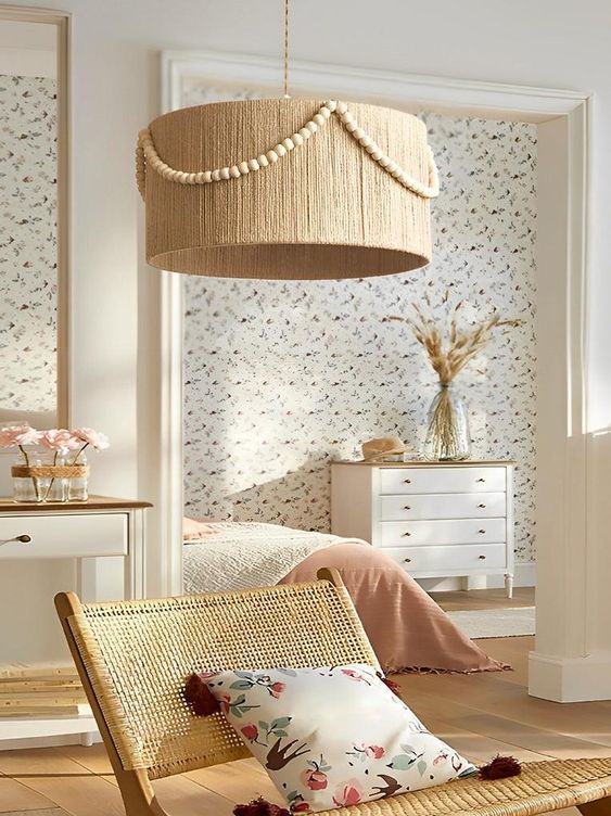 Cozy bedroom with woven lampshade, floral wallpaper, wicker chair, floral pillow, and dresser with dried flowers in vase.