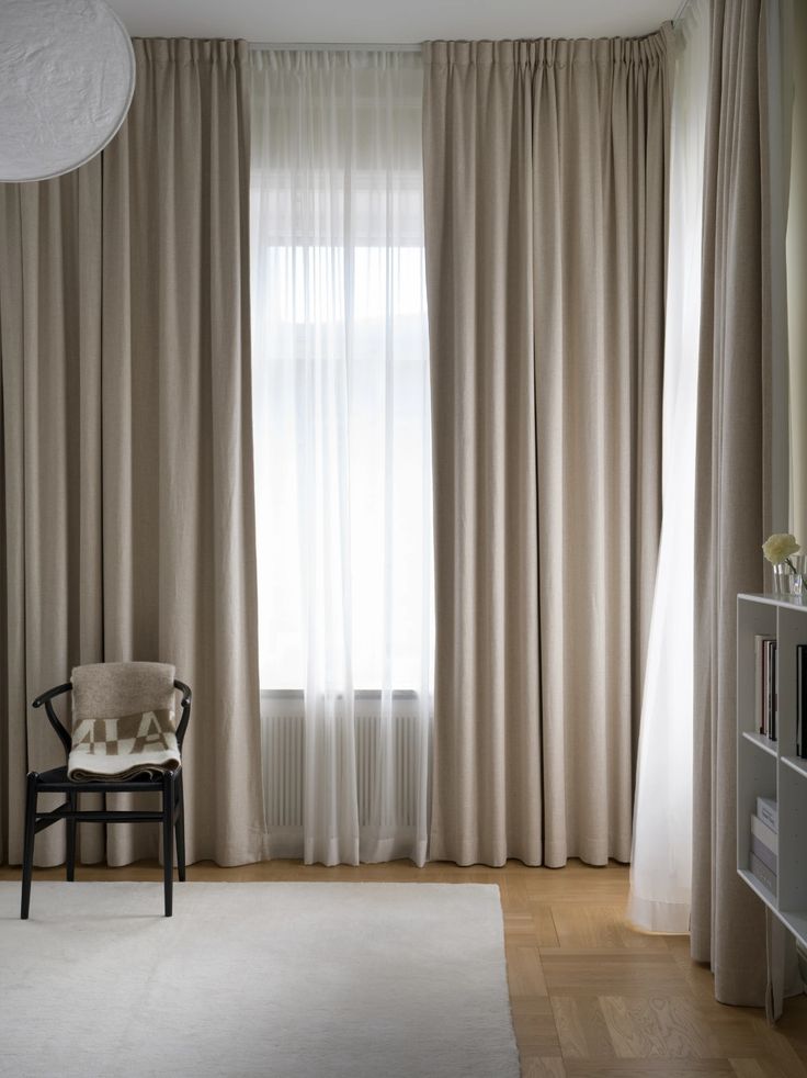 Modern living room with beige floor-length curtains, sheer white drapes, black chair with blanket, and light wood floor.