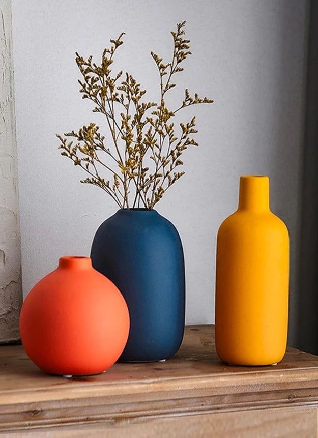 Three colorful vases on wooden table: round orange, tall blue with branches, and yellow bottle-shaped vase, minimalist decor.