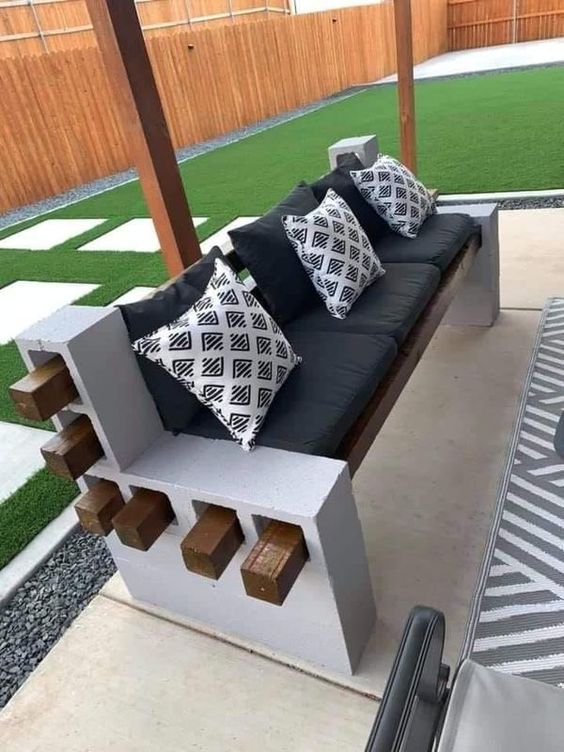 Outdoor cinder block bench with wooden planks, black cushions, and patterned throw pillows on a patio with artificial grass.