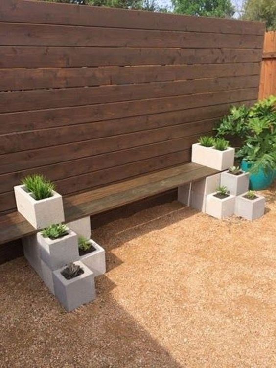 DIY garden bench made from wooden plank and cinder blocks, adorned with small planters in a backyard against a wooden fence.