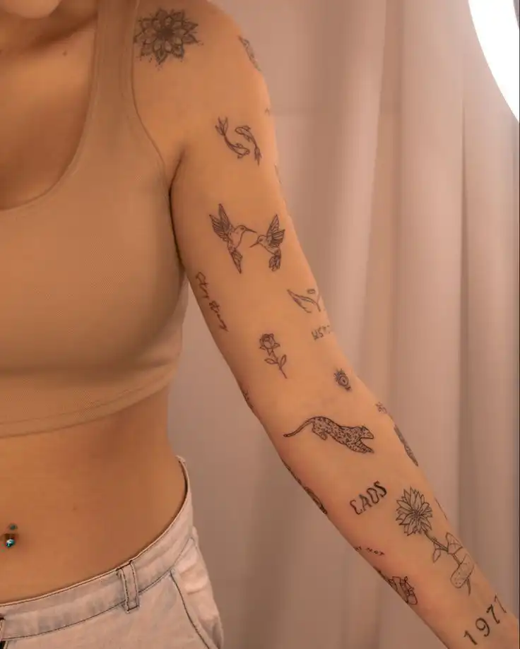 Person with floral and animal tattoos on arm, showcasing creative body art in soft lighting. Wearing a beige crop top and light jeans.