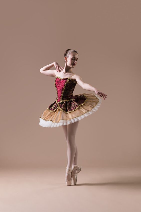 Ballet dancer performing on pointe in a red and gold tutu against a neutral background.