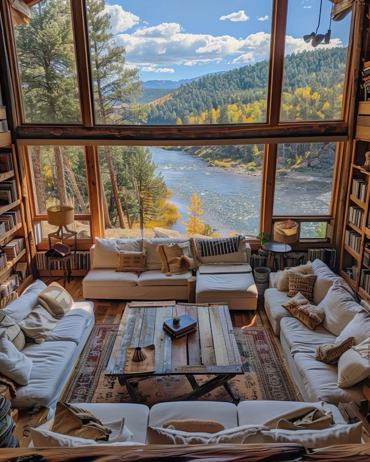 Cozy cabin living room with large windows, scenic river, forest views, plush sofas, and rustic wooden coffee table.