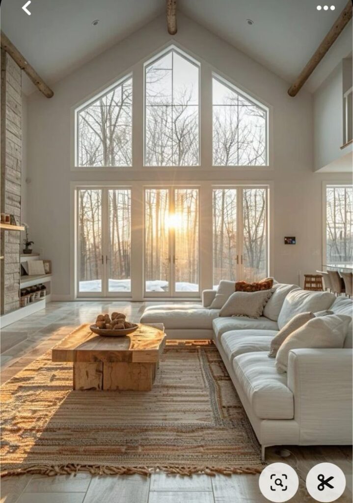 Modern living room with large windows, white sofa, rustic wooden coffee table, and a sunset view through trees.