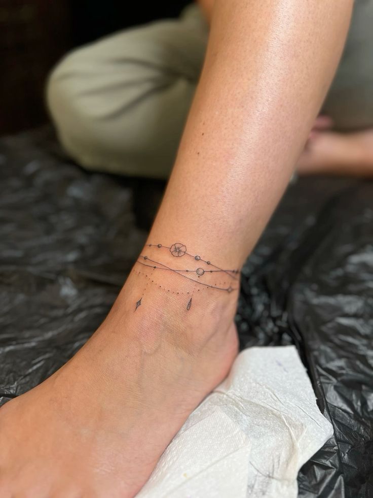 Ankle tattoo with delicate lines, dots, and symbols, including a flower and teardrop, on the left leg.