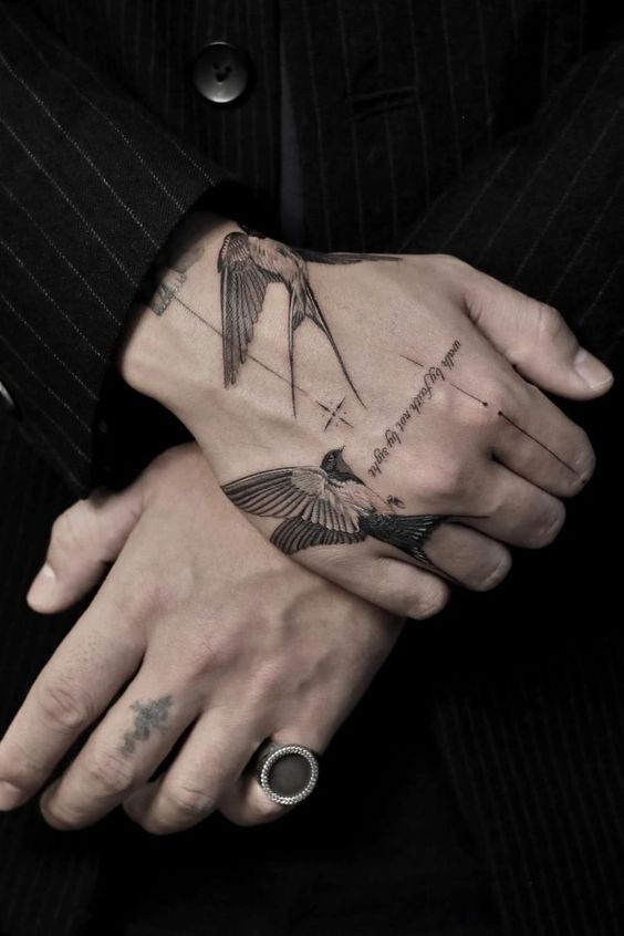 Close-up of crossed hands featuring detailed swallow bird tattoos and a ring on the pinky finger, dressed in a pinstripe suit.