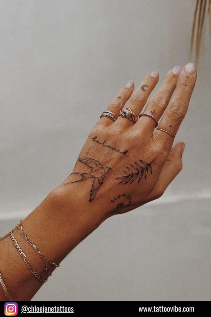 Hand with intricate tattoos of a bird, leaf, and text, adorned with rings and bracelets. Follow @chloejanetattoos on Instagram.