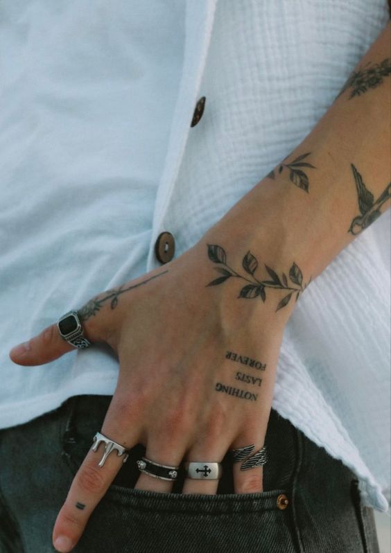 Close-up of a hand with leafy tattoos, wearing multiple rings, resting in a pocket of black jeans.